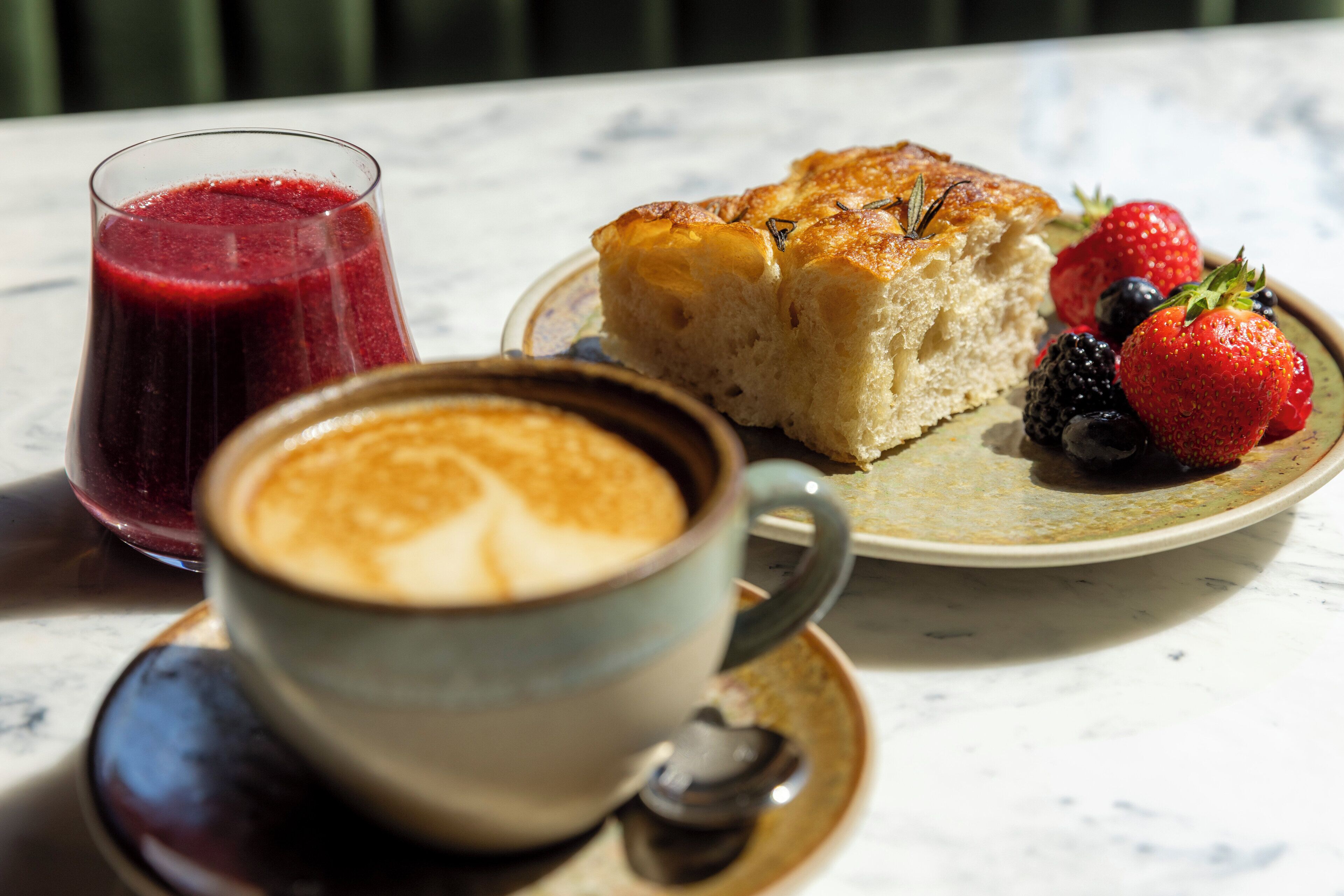 En desserttallerken med et stykke kage og friske bær, et glas rød saft og en kop kaffe på et bord.