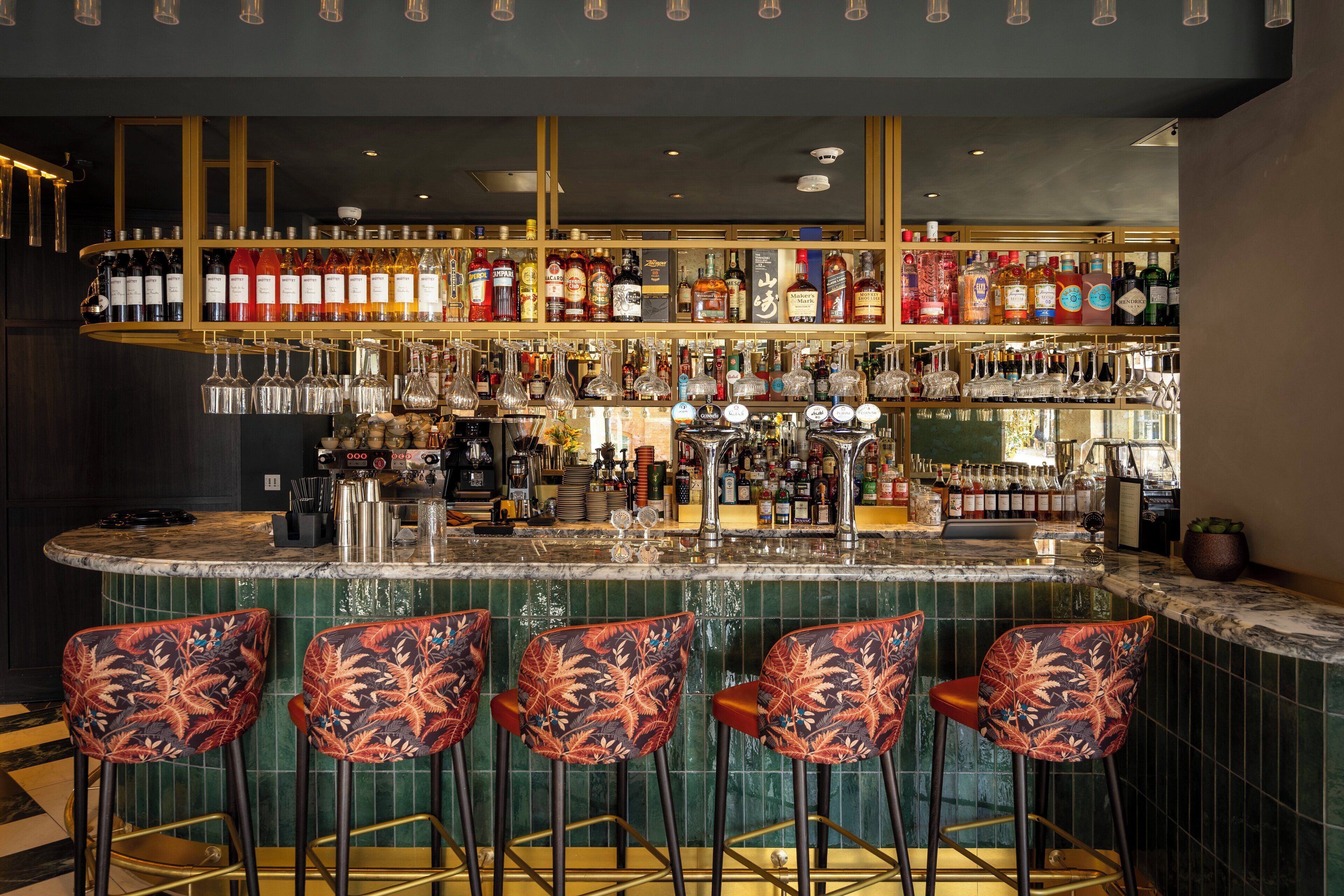Un espace bar doté d’un comptoir en marbre, de nombreuses bouteilles d’alcool et de quatre tabourets de bar à motifs.