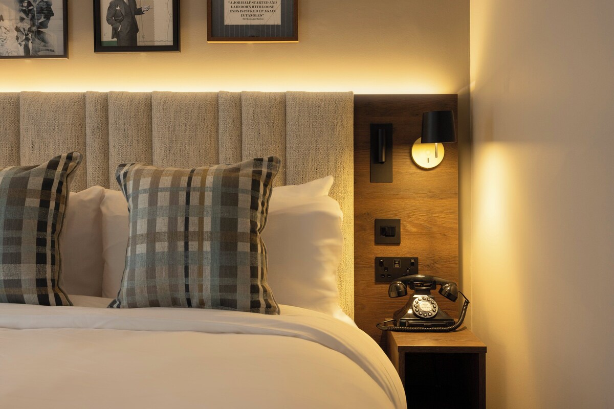 Una cama bien hecha con almohadones escoceses, una mesita de noche de madera con un teléfono antiguo y cuadros en la pared.