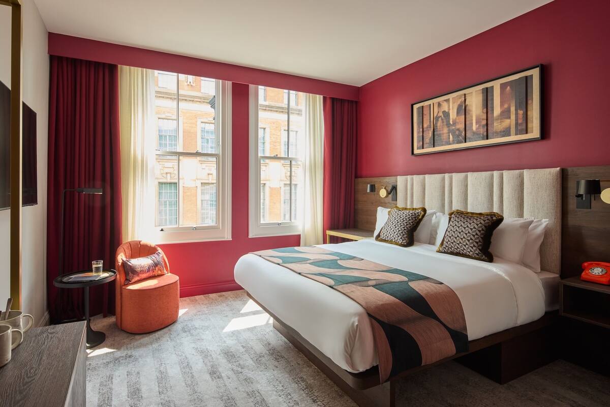 Ein Hotelzimmer mit einem großen Bett, einer roten Akzentwand, einem Gemälde und Blick auf Gebäude durch die Fenster.