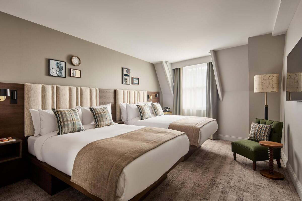 Ein Hotelzimmer mit zwei Betten, einem grünen Sessel, einem runden Tisch und einem großen Fenster mit Vorhängen.