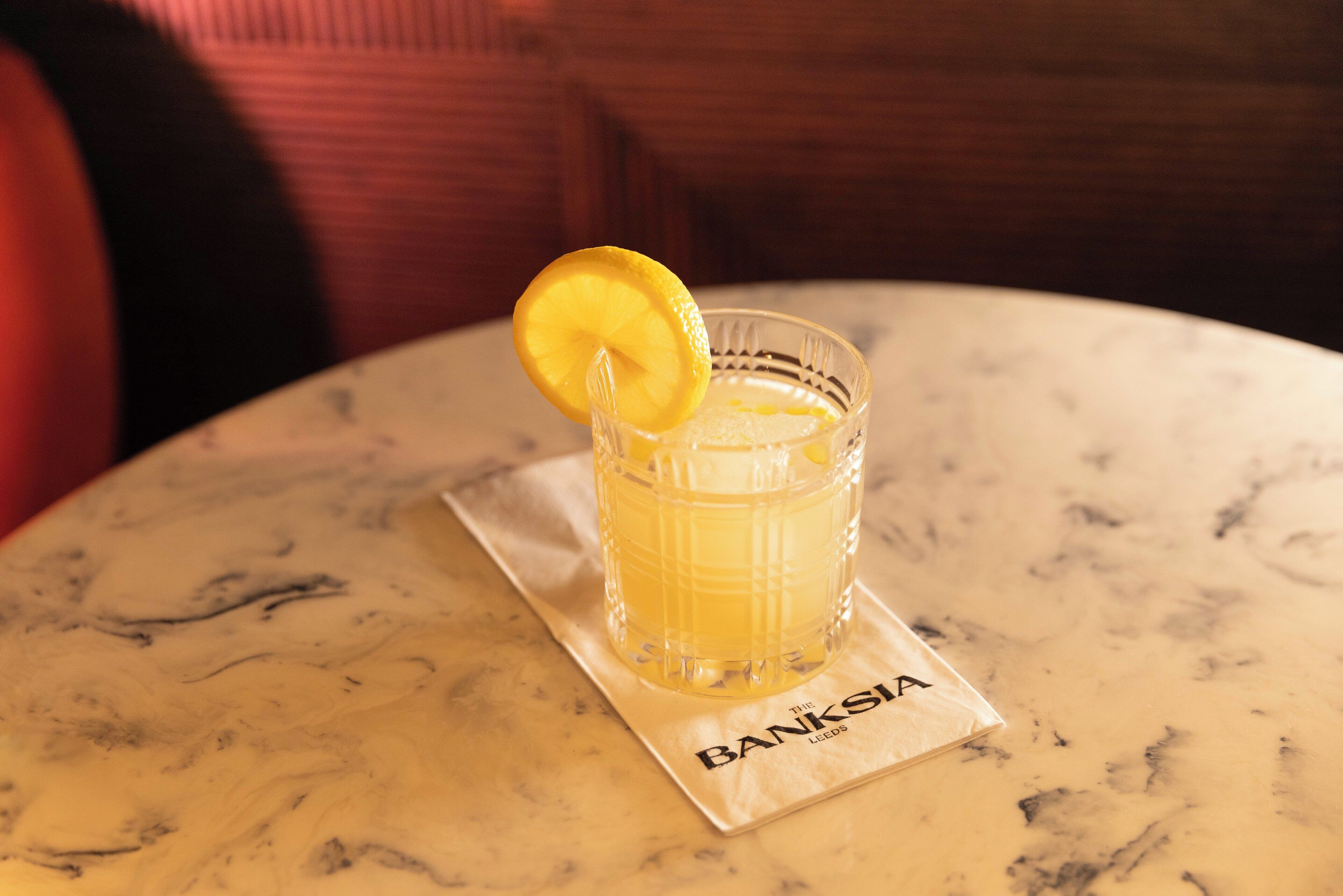 A glass of a yellow beverage with a lemon slice on a marble table with a napkin labeled "BANKSIA".