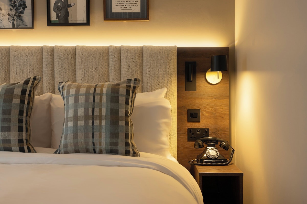 Una cama bien hecha con almohadones escoceses, una mesita de noche de madera con un teléfono antiguo y cuadros en la pared.