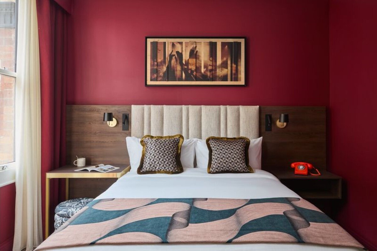 Habitación de hotel con una cama grande, una mesita de noche con teléfono, un cuadro enmarcado en la pared y una ventana con cortinas.