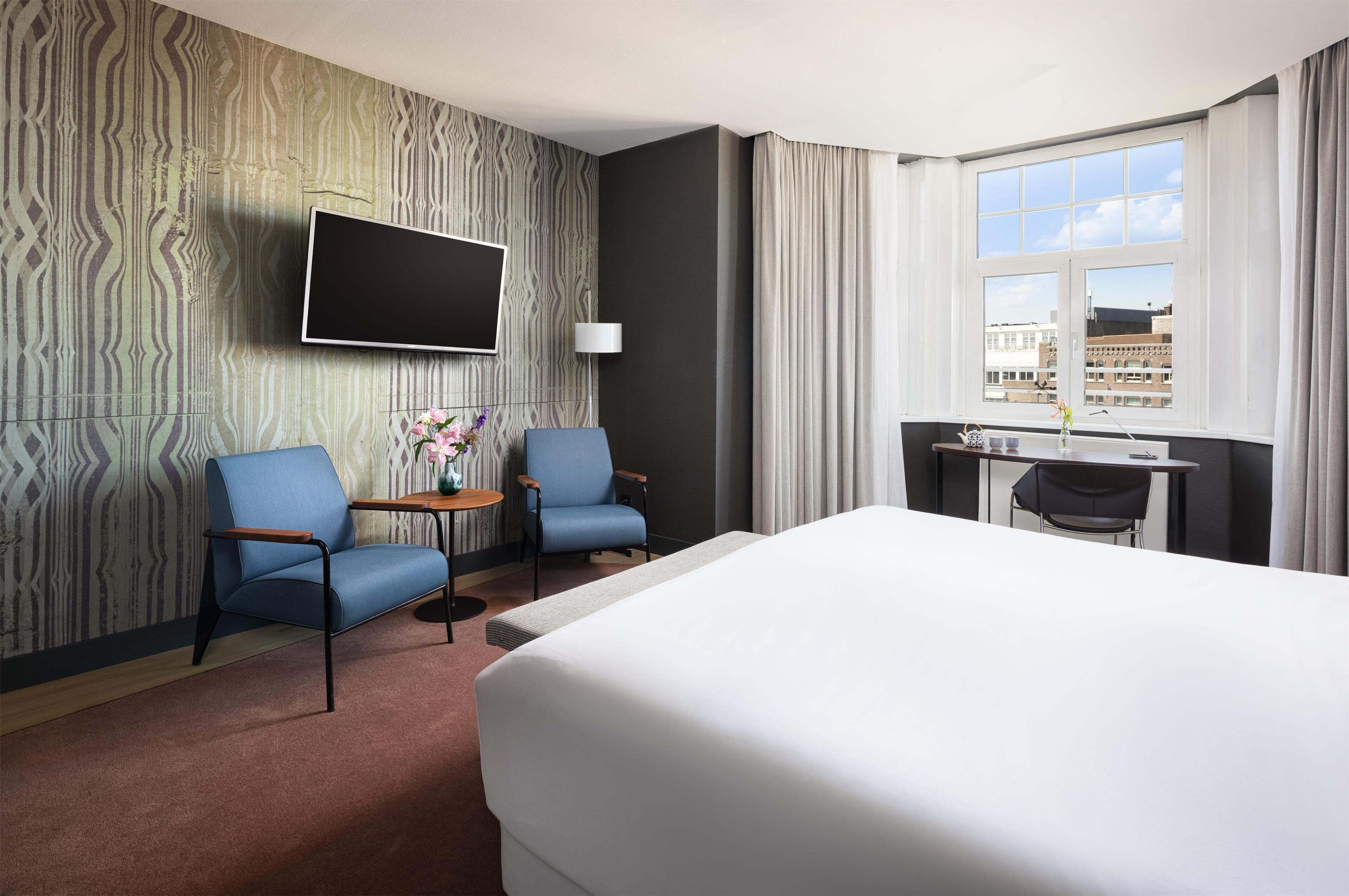 Une chambre d’hôtel avec un grand lit, deux fauteuils, une petite table ornée d’un vase de fleurs, un téléviseur fixé au mur et une fenêtre avec des rideaux.