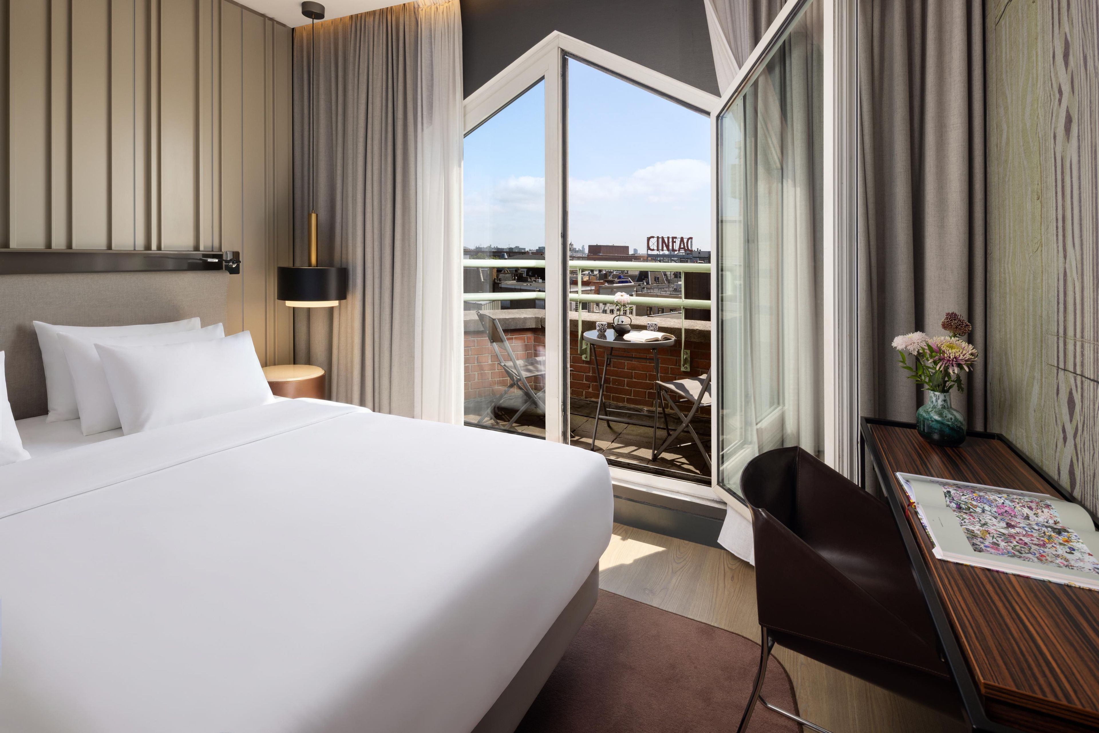 Une chambre d’hôtel dotée d’un grand lit, d’un bureau avec une chaise, d’une fenêtre donnant sur la ville et d’un balcon avec des sièges extérieurs.
