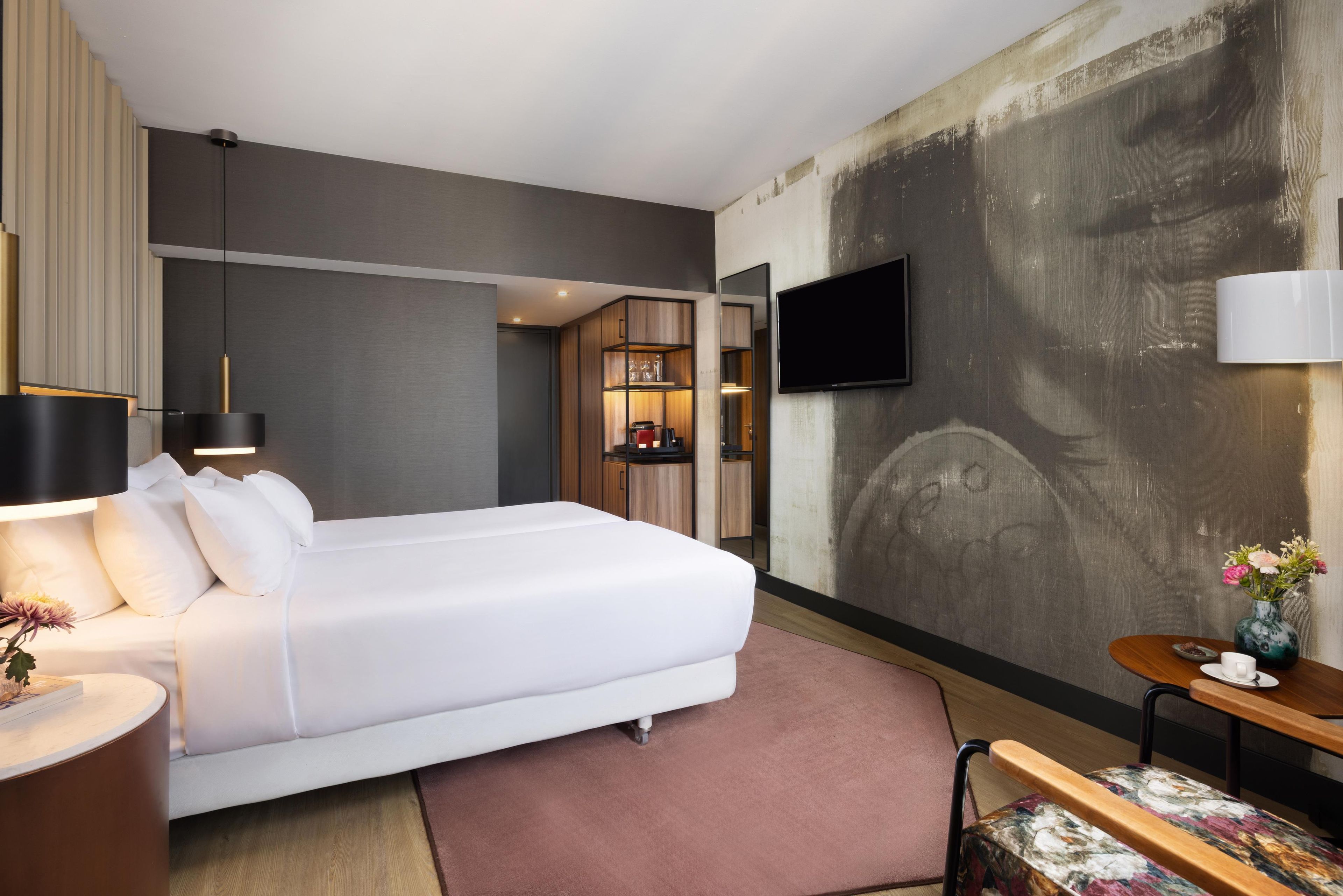 Una habitación de hotel moderna con una cama grande, un televisor de pantalla plana, un escritorio con una silla y una mesita con un jarrón de flores.