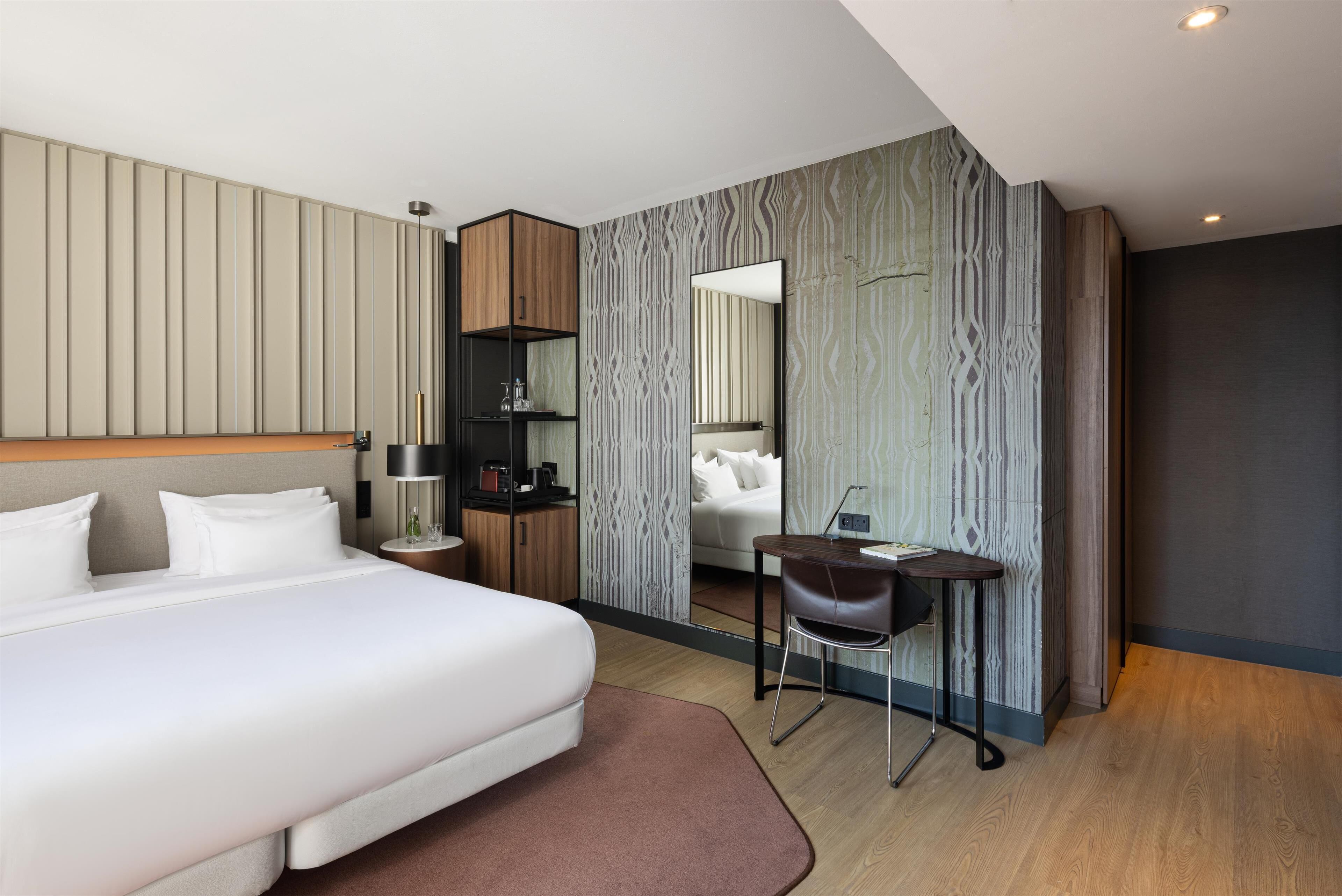 A modern hotel room with a large bed, a desk, a chair, a wardrobe, and a mirror.