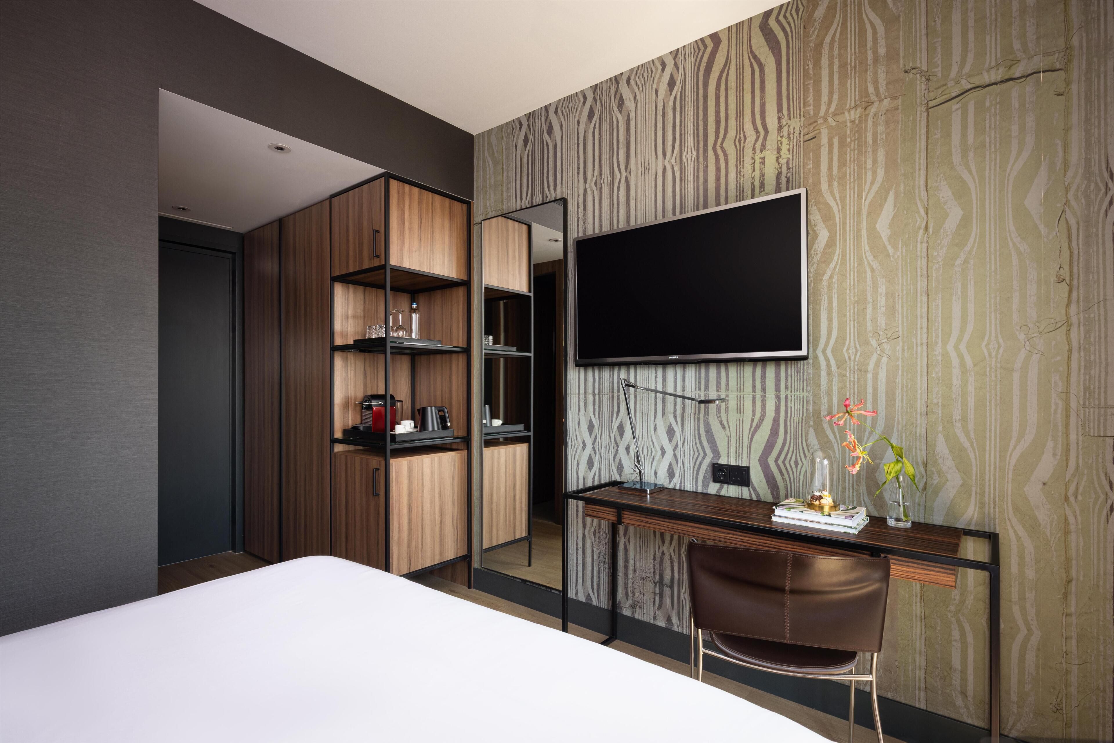 Quarto de hotel moderno com uma cama grande, uma escrivaninha com cadeira, televisão de tela plana e um guarda-roupa com prateleiras.
