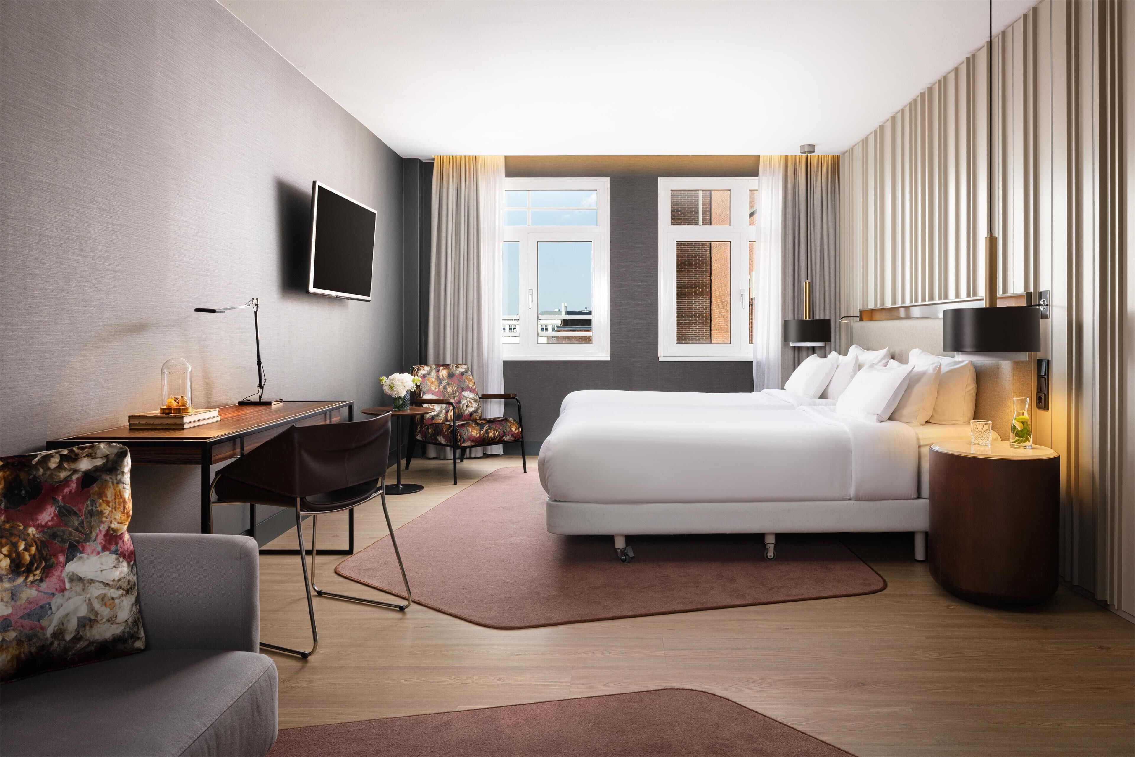 A modern hotel room with a bed, desk, chair, TV, and a view of the city.