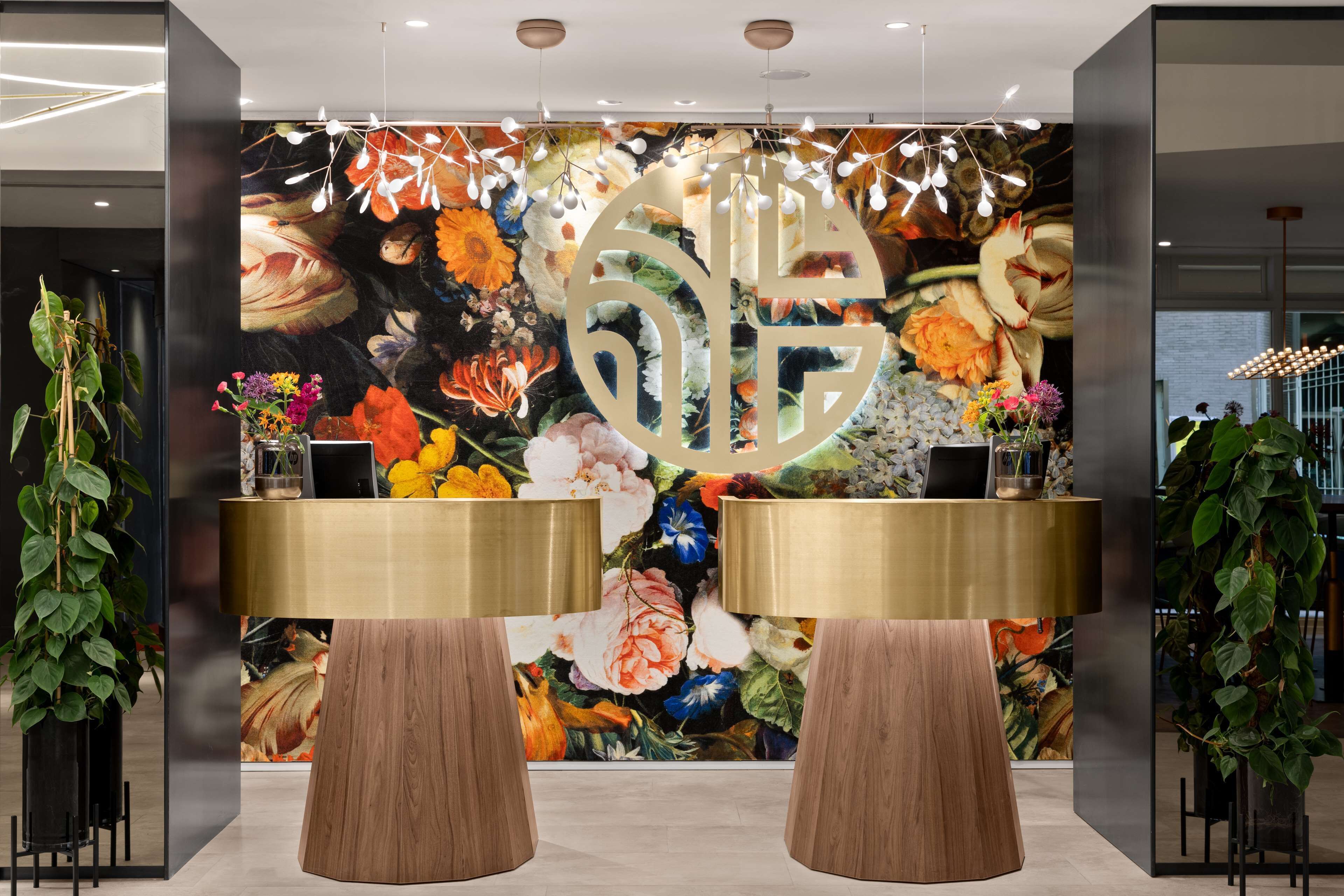 Un área de recepción con un mural floral, dos escritorios con marcos dorados y grandes macetas.