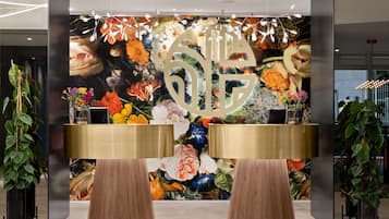 Un área de recepción con un mural floral, dos escritorios con marcos dorados y grandes macetas.