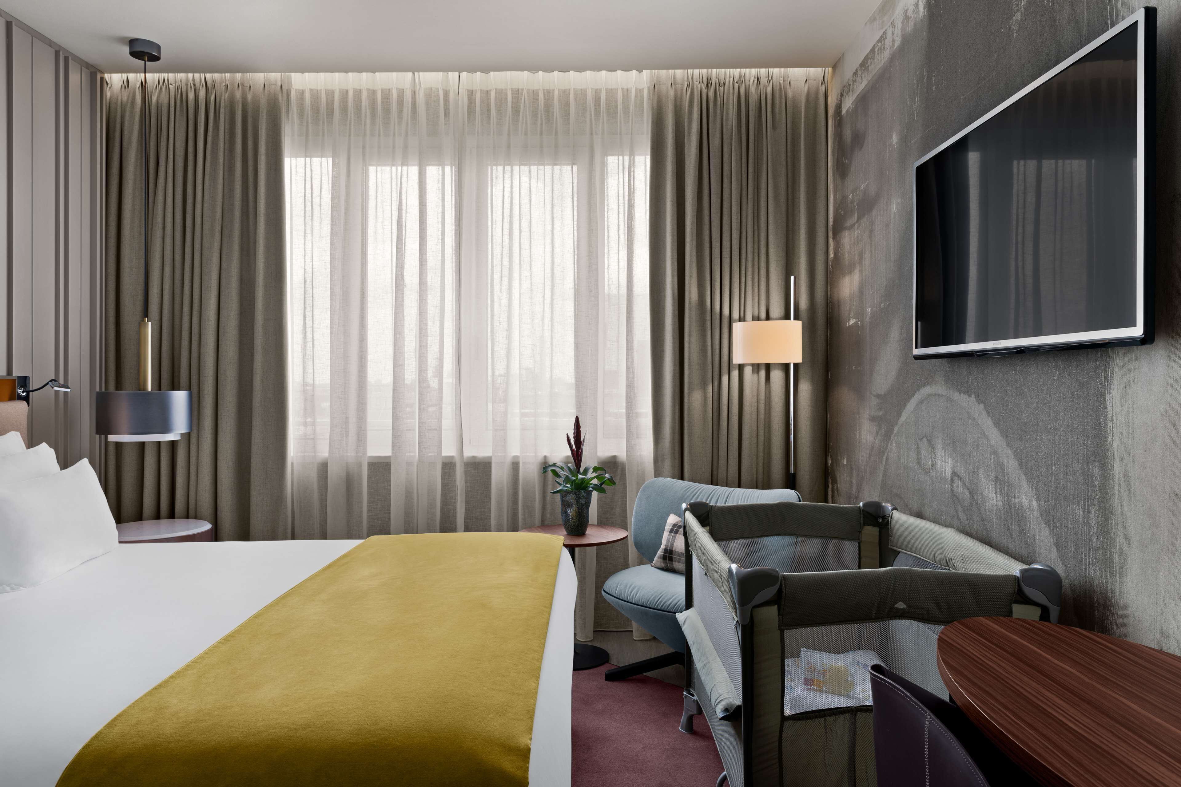 Une chambre d’hôtel avec un grand lit, un bureau, une chaise, un téléviseur fixé au mur et une fenêtre avec des rideaux.