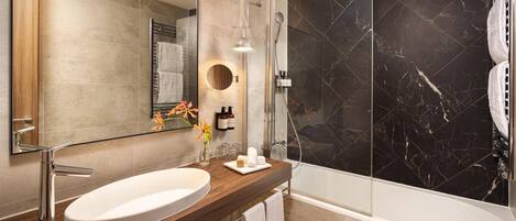A modern bathroom with a large mirror, a wooden vanity, a white sink, a shower with black marble tiles, and a towel rack.