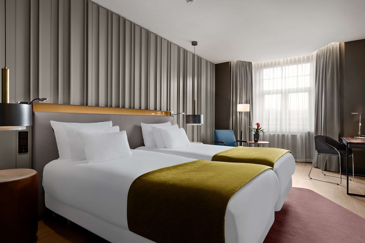 A hotel room with two beds, a desk, a chair, a lamp, and a view of the outdoors.