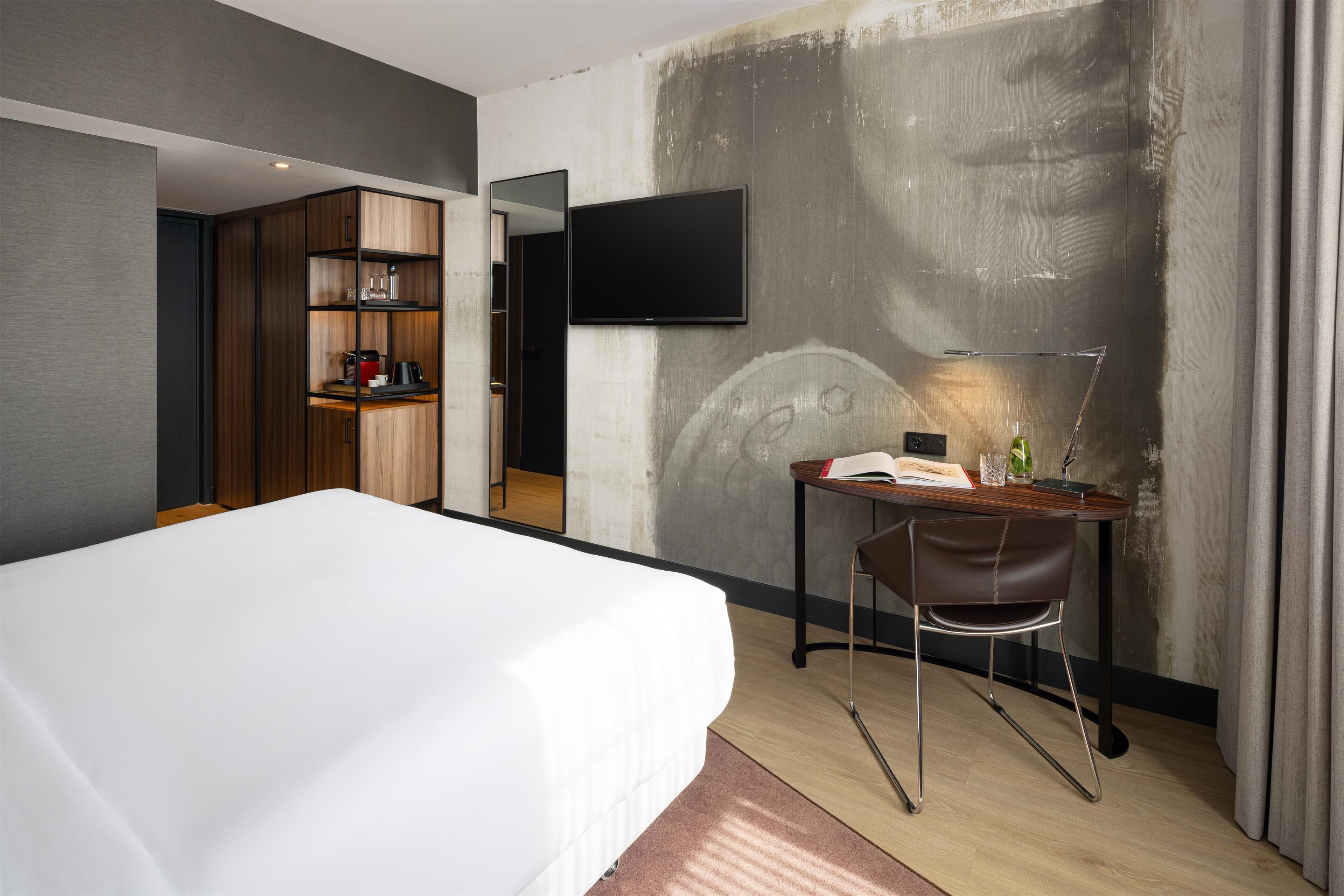 Una habitación de hotel moderna con una cama grande, un escritorio con silla, un televisor de pantalla plana y un armario con estantes.