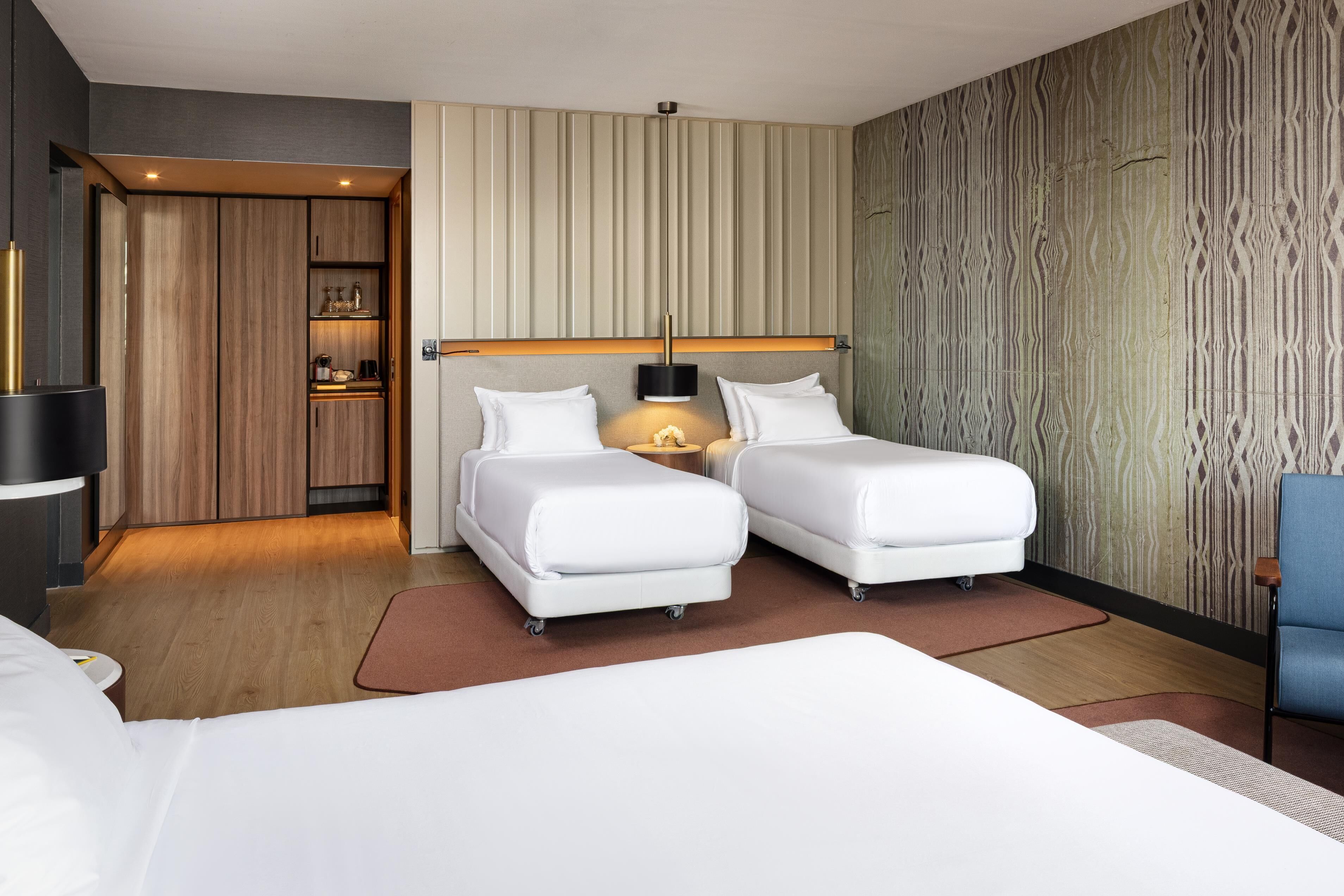 A modern hotel room with two single beds, a small kitchenette, and a large window.