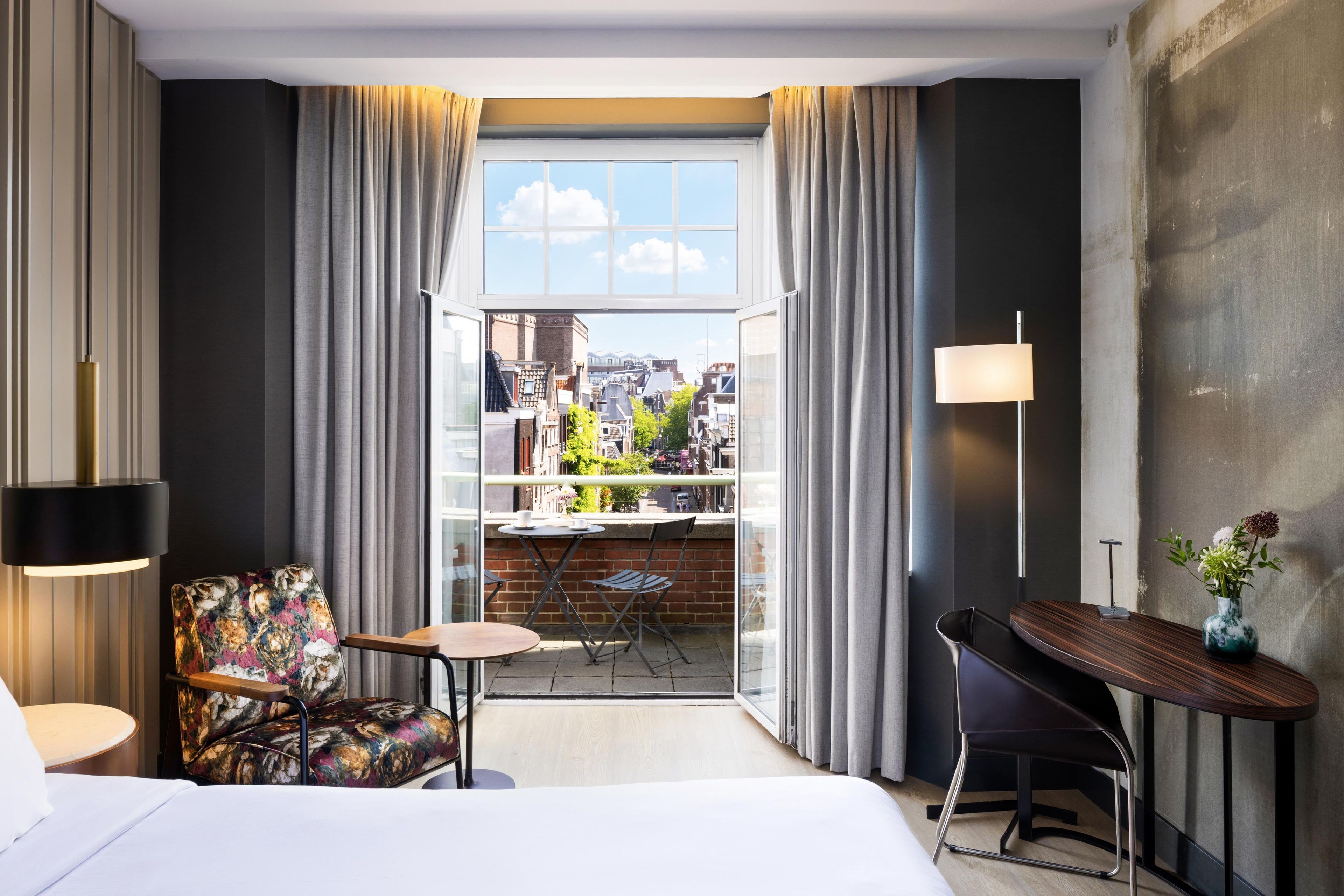 Una habitación de hotel con balcón, una cama, un escritorio, una silla y una lámpara.