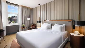 Habitación de hotel moderna con una cama grande, una mesita de noche con lámpara, una silla y vistas a la ciudad a través de una ventana.