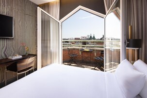 A hotel room with a large bed, a desk with a chair, a TV, and a balcony with a view of the city.
