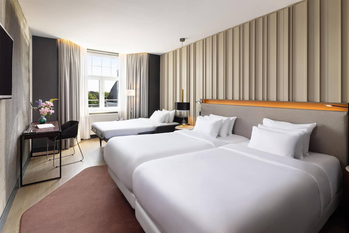A hotel room with two beds, a desk, a chair, a TV, and a window with curtains.