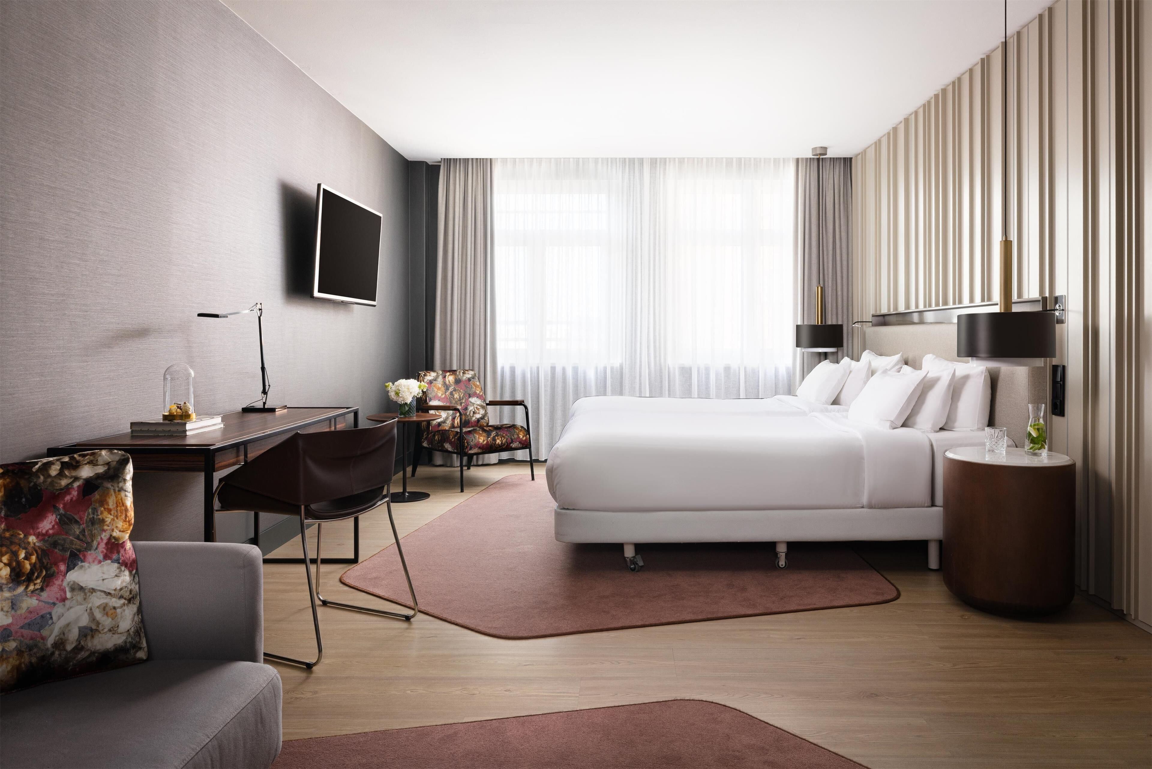 Una habitación de hotel moderna con una cama grande, un escritorio, una silla, un televisor y una mesita de noche con una lámpara.