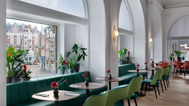 Un restaurante con asientos tipo banca verdes y mesas redondas, ventanas amplias con vista a la ciudad y plantas en macetas.