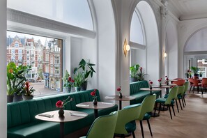 Un restaurant avec des banquettes vertes et des tables rondes, de grandes fenêtres donnant sur la ville et des plantes en pot.