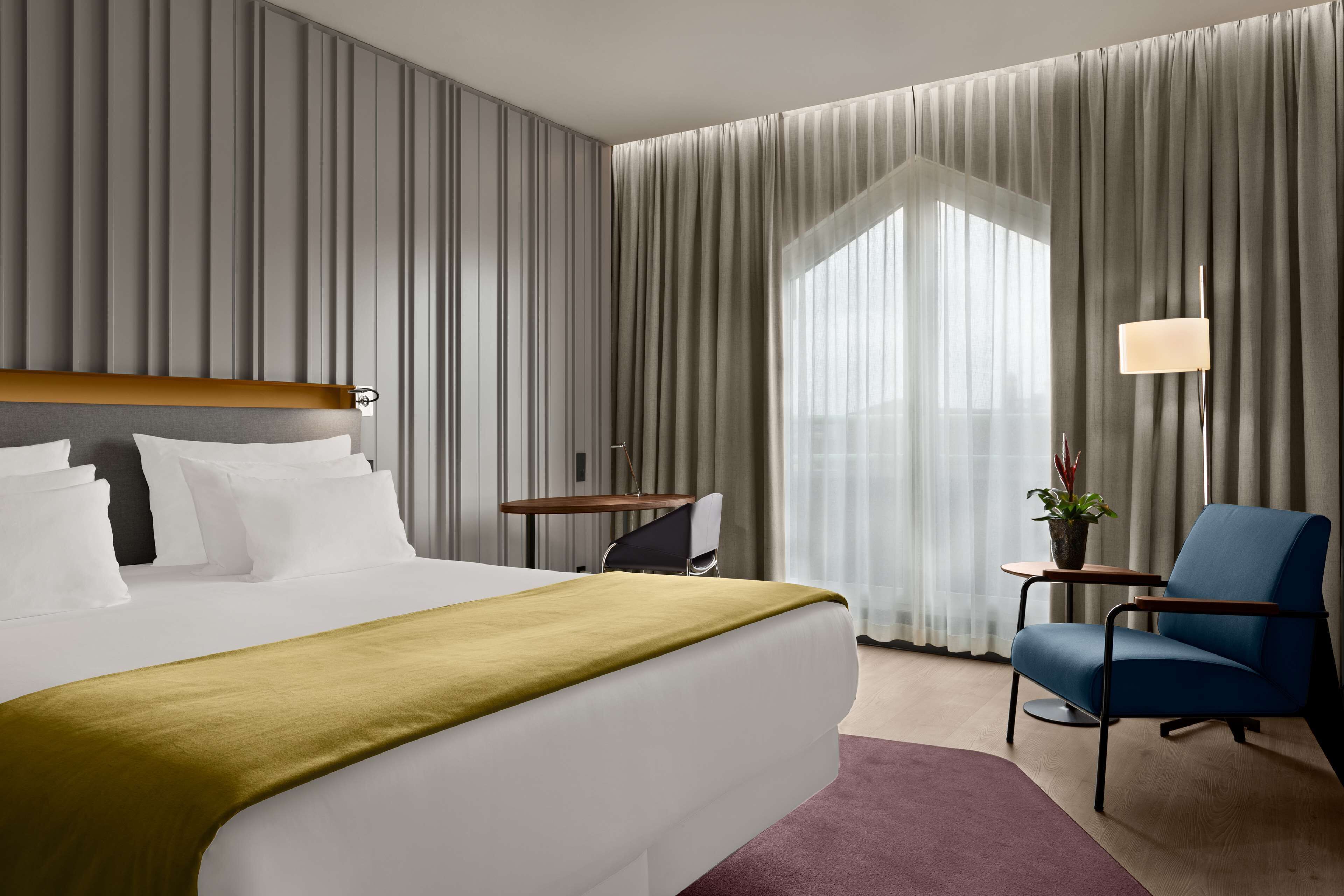 A hotel room with a large bed, a desk, a chair, and a view through sheer curtains.