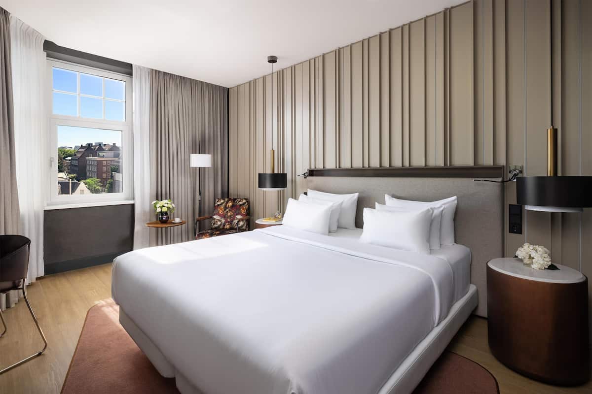 A modern hotel room with a large bed, a view of the city, and a small side table with a lamp.