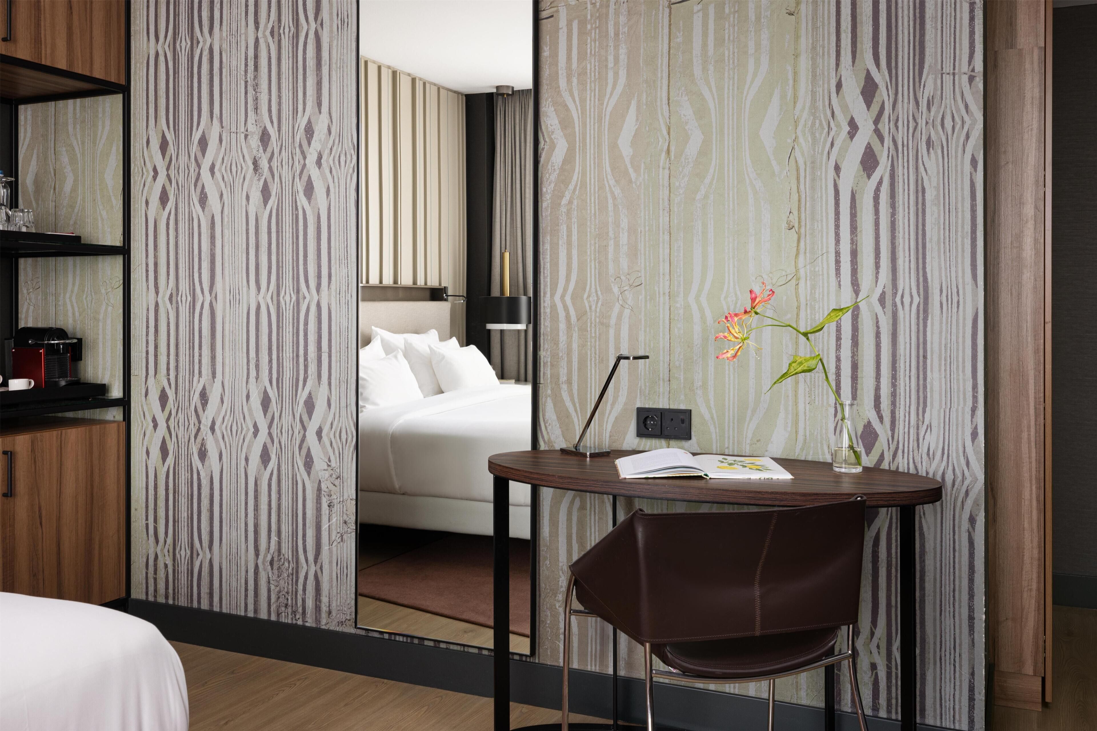 A hotel room with a bed, a dining table with a chair, and a small table with a vase of flowers.