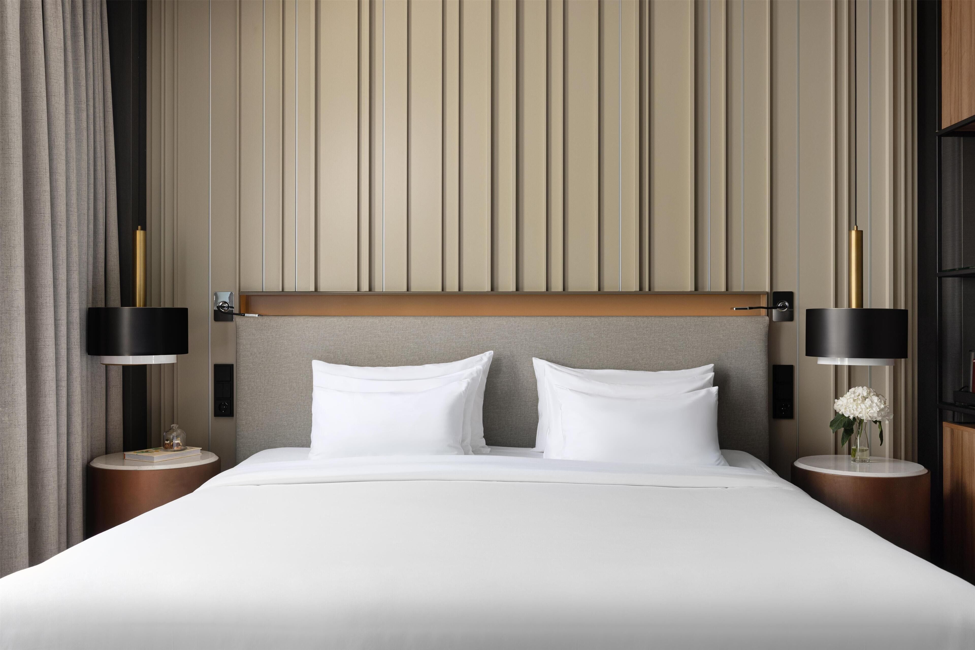 Una habitación de hotel con una cama grande, dos mesitas de noche, un cabecero moderno y una pared con líneas verticales.