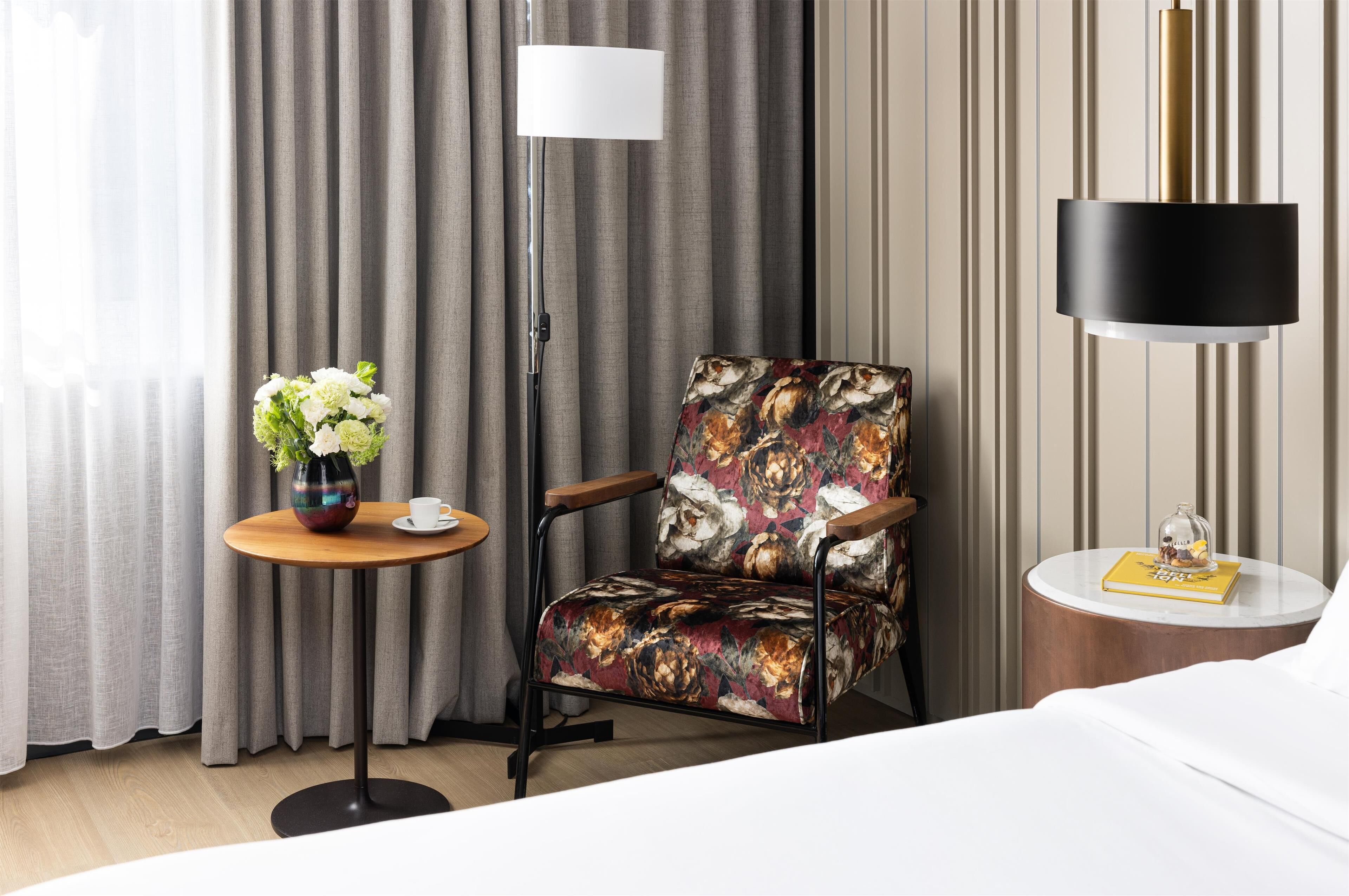 Habitación de hotel con una cama, una silla de estampado floral, una mesita redonda con un jarrón de flores, una lámpara de pie y una mesita de noche con un libro y un objeto decorativo.