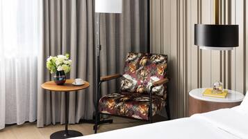 Habitación de hotel con una cama, una silla de estampado floral, una mesita redonda con un jarrón de flores, una lámpara de pie y una mesita de noche con un libro y un objeto decorativo.