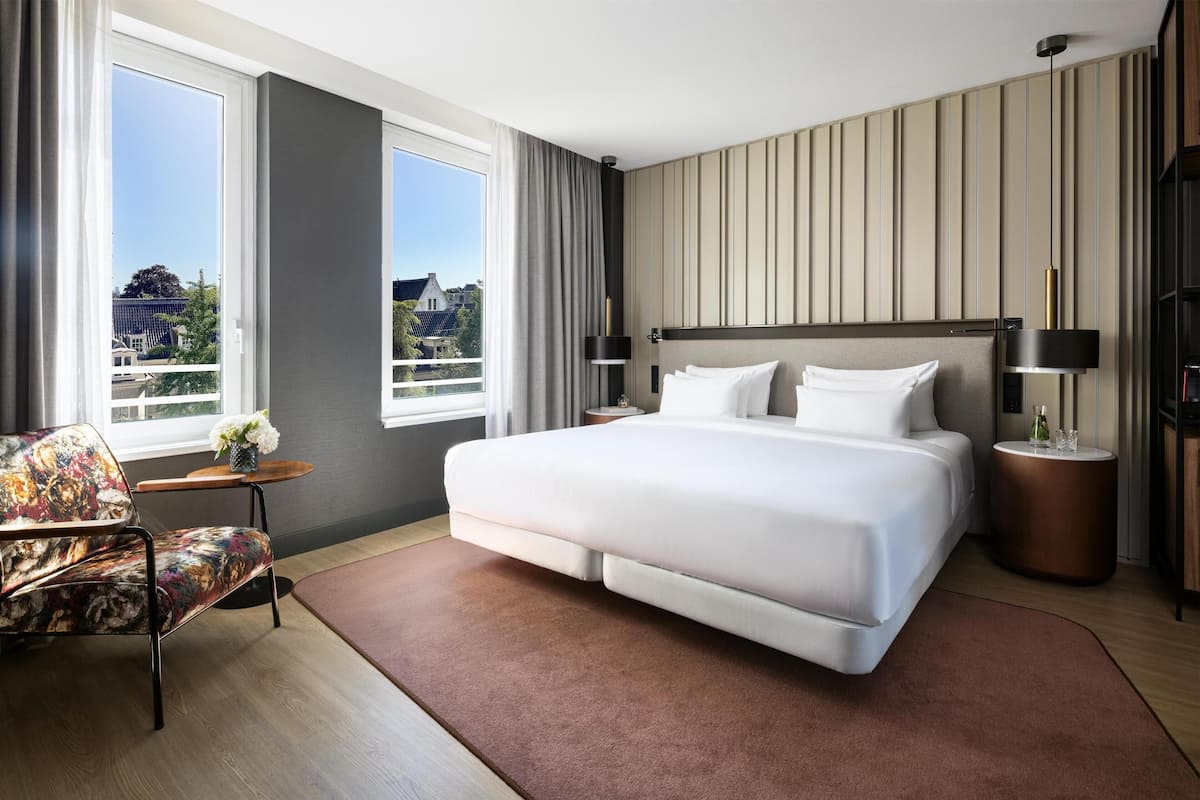 A modern hotel room with a large bed, a chair, a small table, and a view of the city.