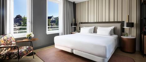 A modern hotel room with a large bed, a chair, a small table, and a view of the city.
