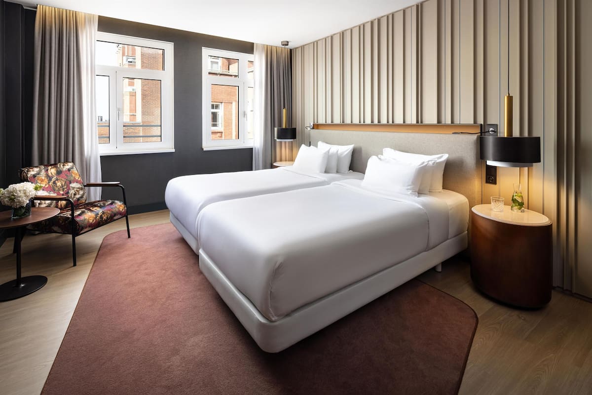 A hotel room with two beds, a round side table, a chair with a floral cushion, a window with curtains, and a wall-mounted lamp.