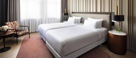 A hotel room with a large bed, a side table, a chair, and a view of the outdoors through the window.