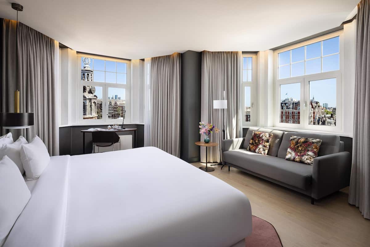 A modern hotel room with a large bed, a sofa, a desk, and a view of the cityscape.