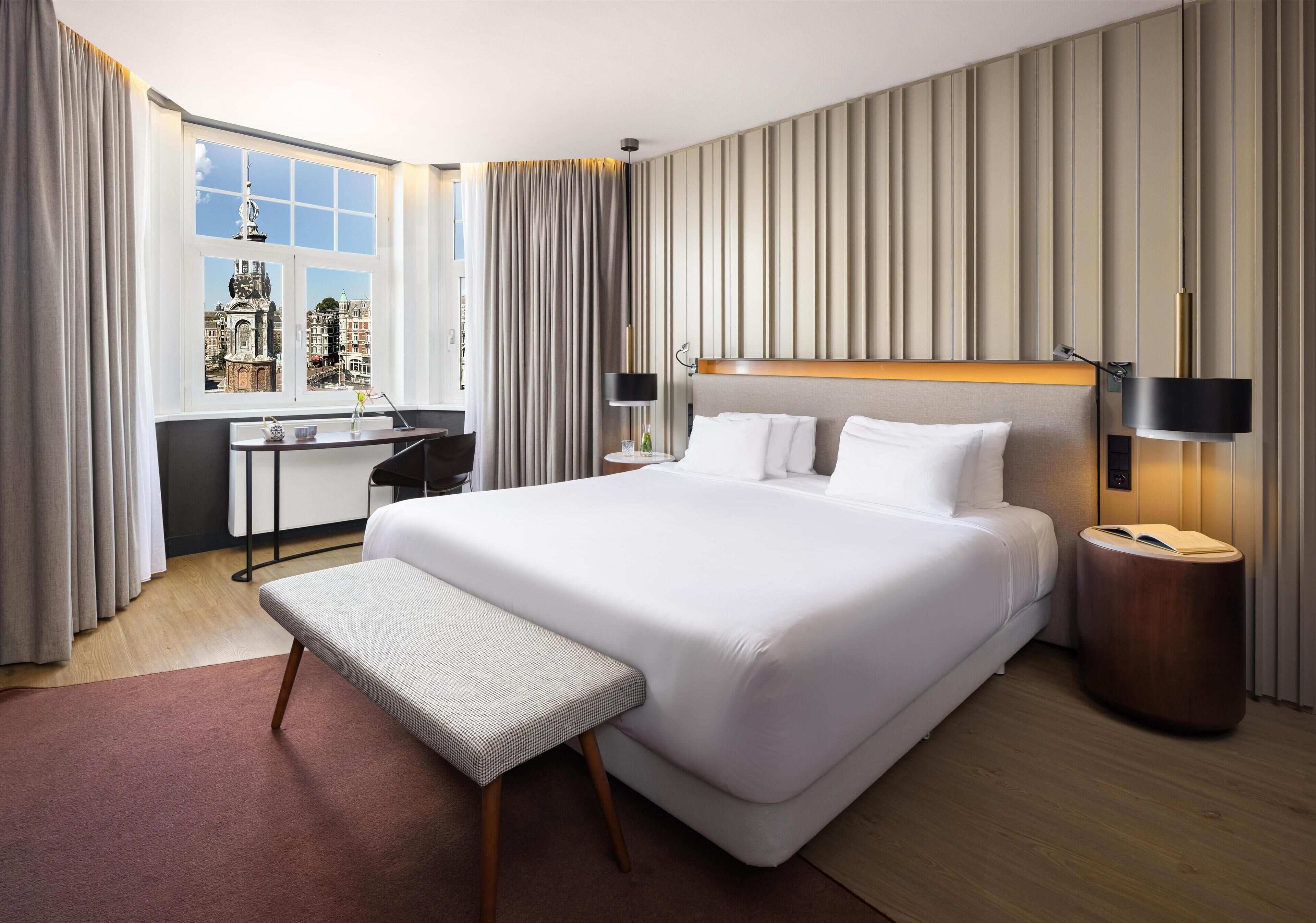 A modern hotel room with a large bed, a desk, and a view of a cityscape through a large window.