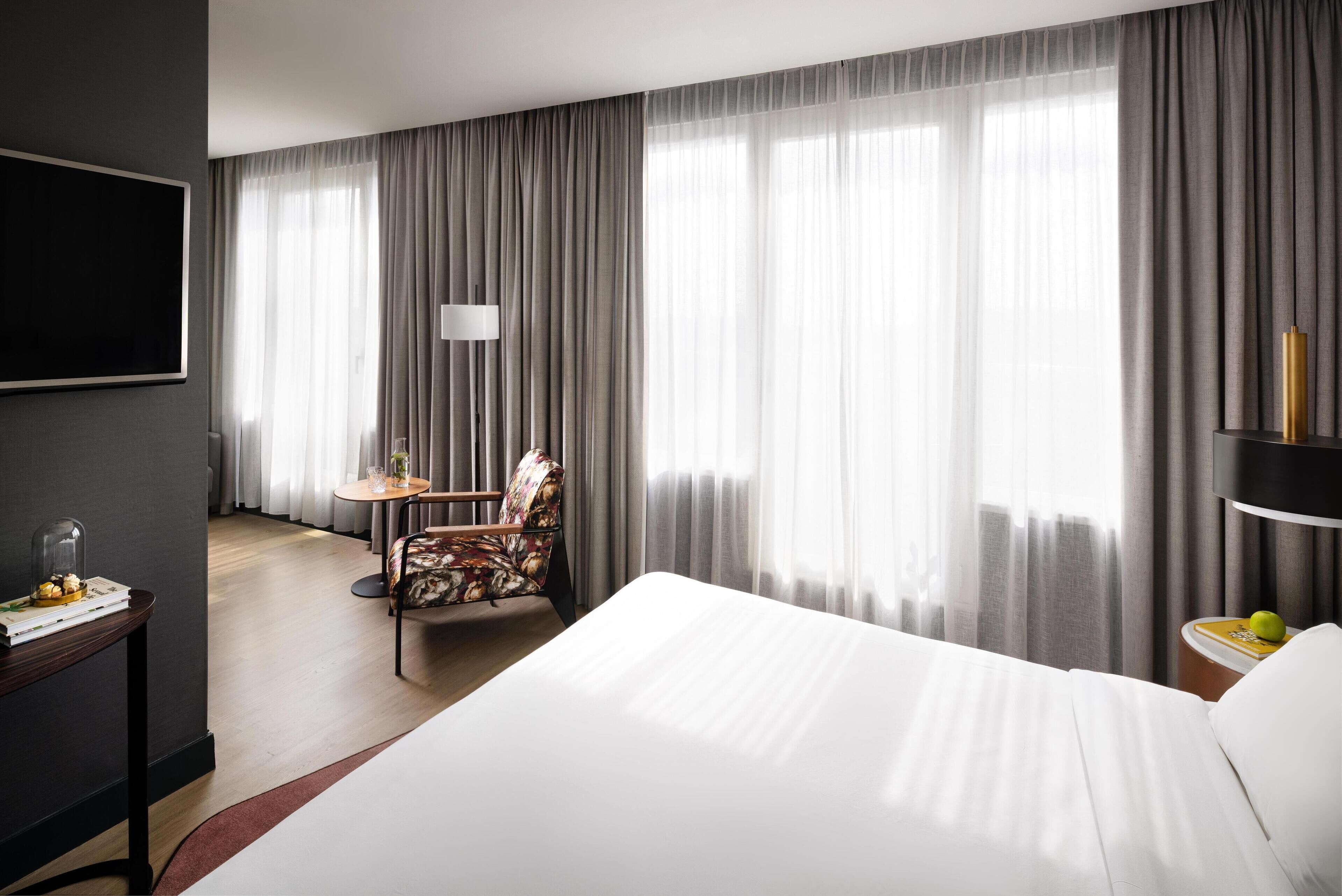 A hotel room with a large bed, a desk with a chair, a TV mounted on the wall, and a window with curtains.