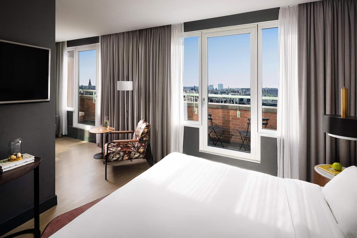 A hotel room with a large bed, a desk with a chair, a small table, and a view of the city through large windows.