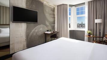 Habitación de hotel con una cama grande, un escritorio, una silla, un televisor montado en la pared y un ventanal con vista a la ciudad.