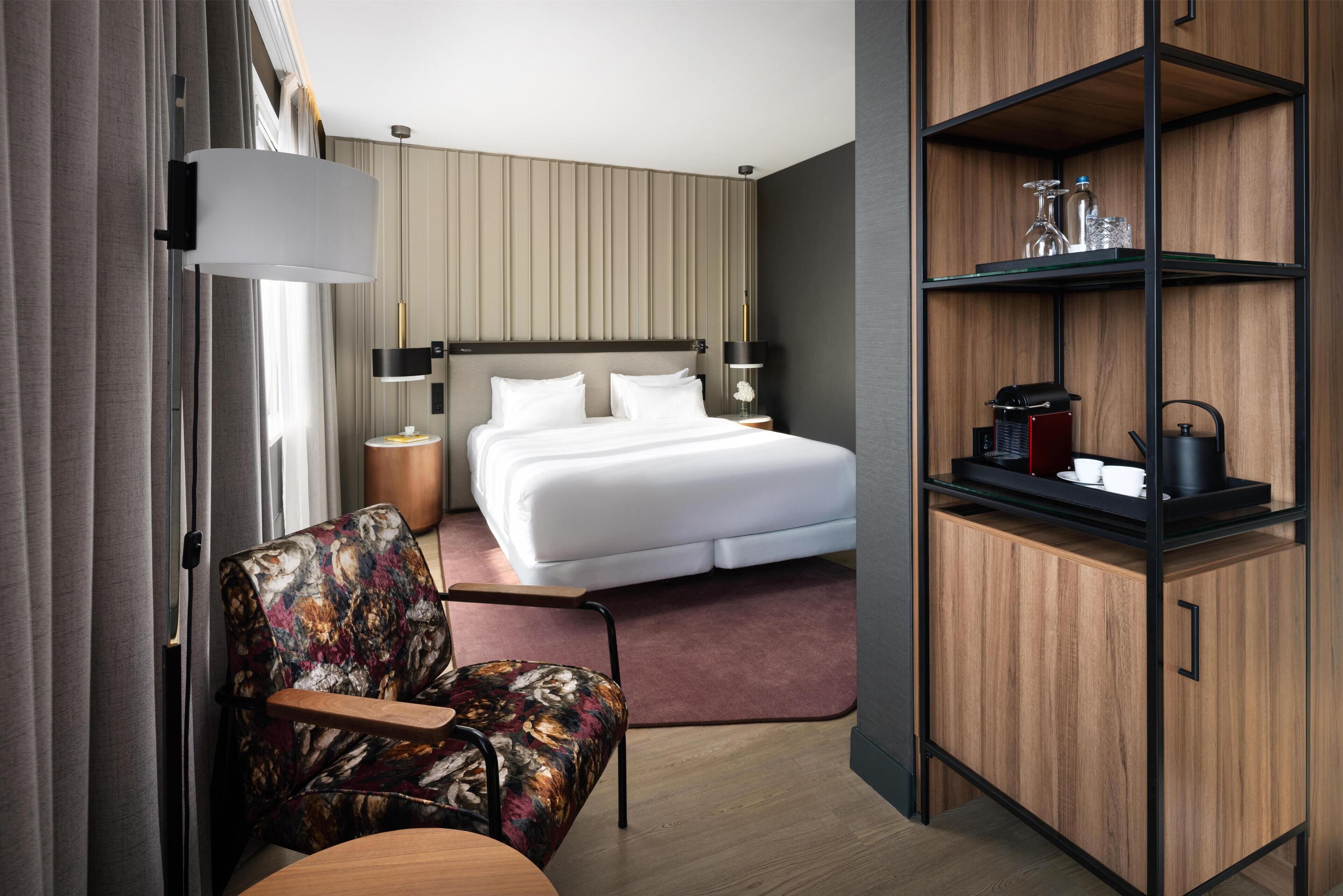 A hotel room with a bed, a chair, a desk, a shelf with items, and a window with curtains.