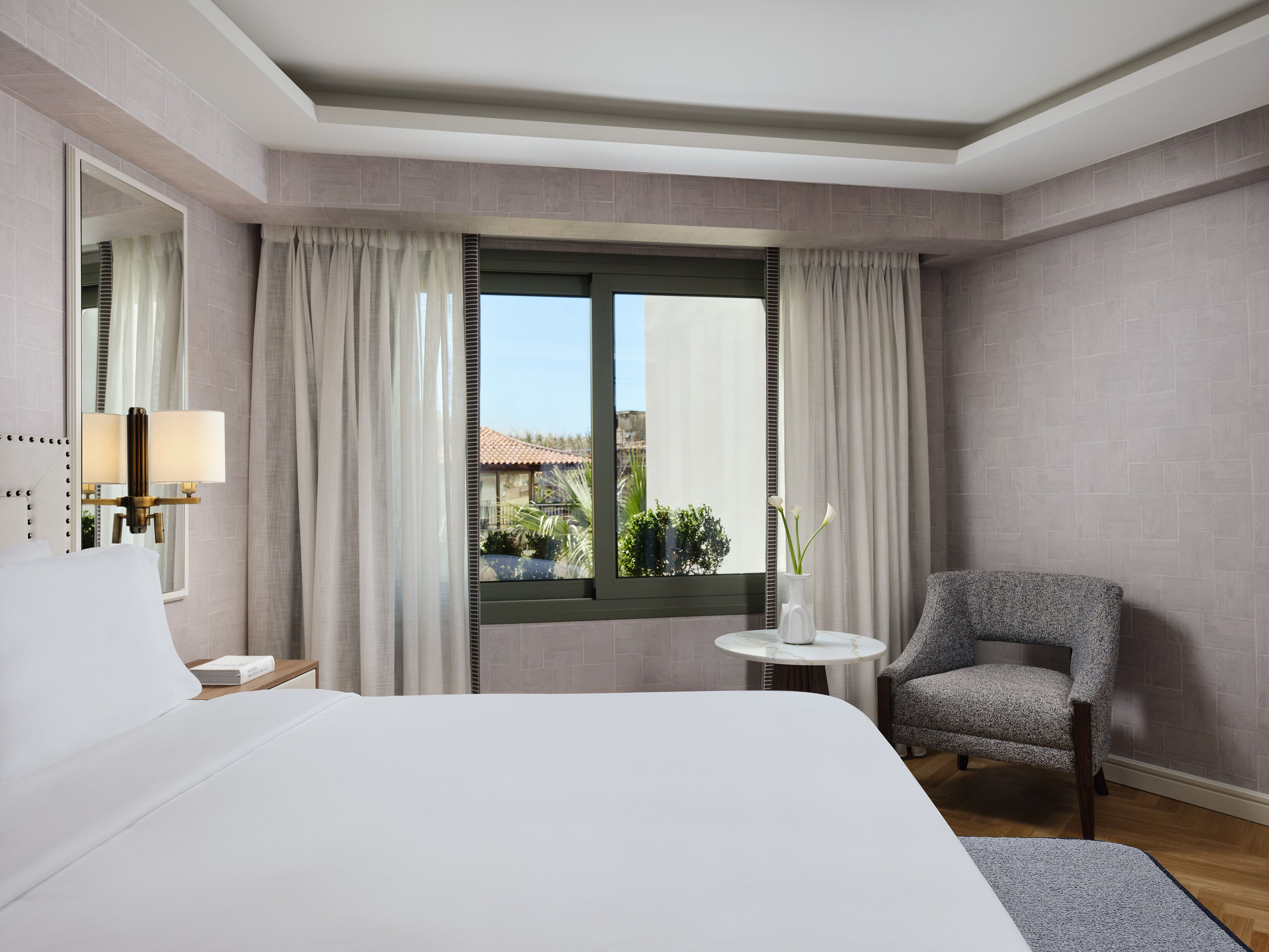 A hotel room with a bed, a chair, a small table with a vase, and a window with curtains.