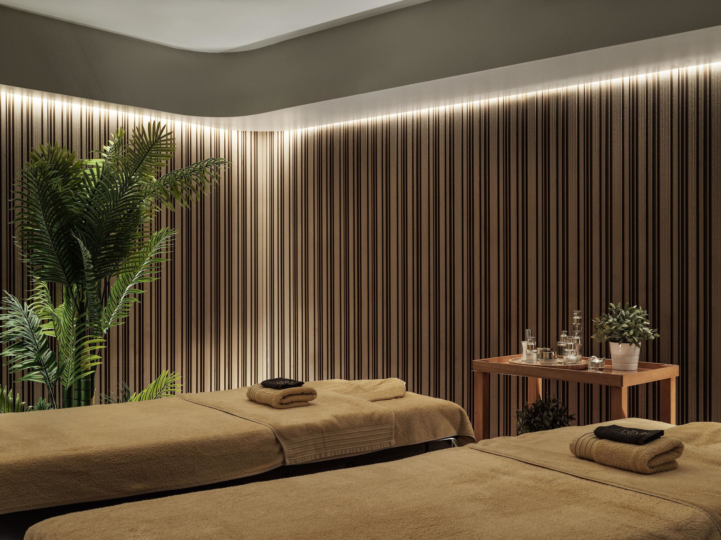 Ein Spa-Raum mit zwei Massagetischen, einem Holztisch mit Pflegeprodukten und einer großen Pflanze.