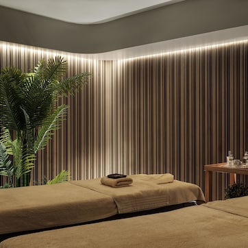 A spa room with two massage tables, a wooden side table with toiletries, and a large plant.