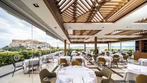 Un restaurante con mesas redondas dispuestas para comer, grandes ventanales que ofrecen vista a una fortaleza histórica y un diseño de techo moderno.