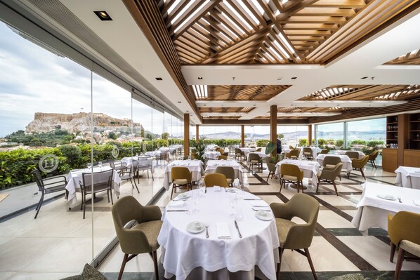 A restaurant with round tables set for dining, large windows offering a view of a historic fort, and a modern ceiling design. - Electra Palace Athens (Athens)