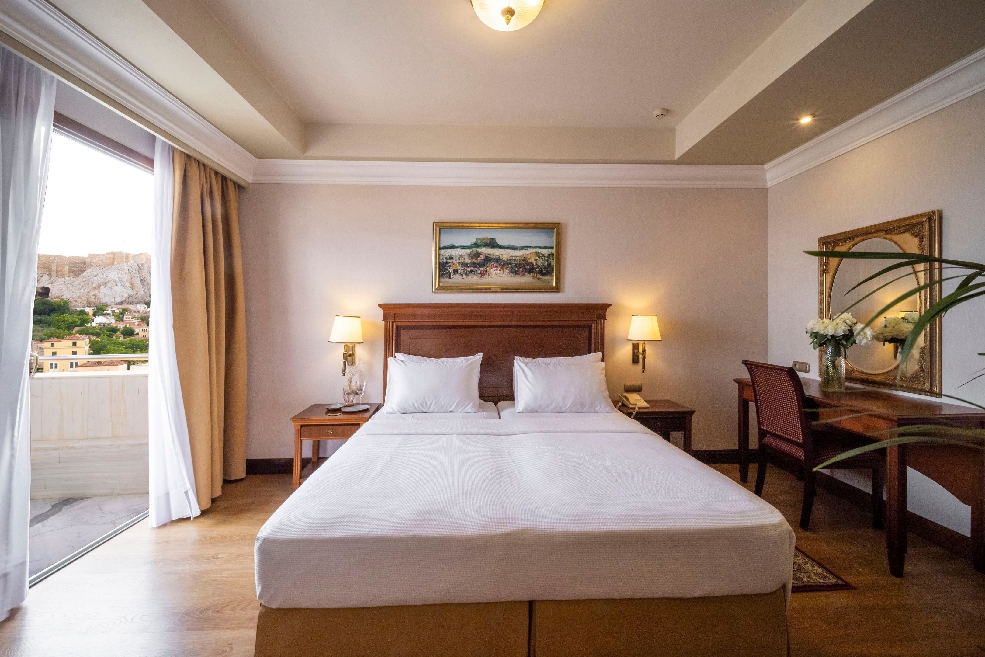Habitación de hotel con una cama grande, dos mesitas de noche, un escritorio con silla, un cuadro en la pared y vistas a la ciudad desde la ventana.